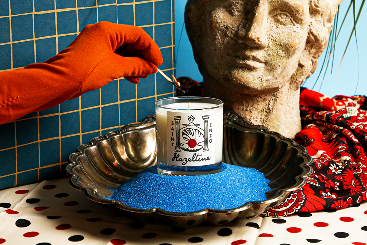 Hand lighting a Hazeltine Saint Enzo candle displayed in a metal dish with blue sand.