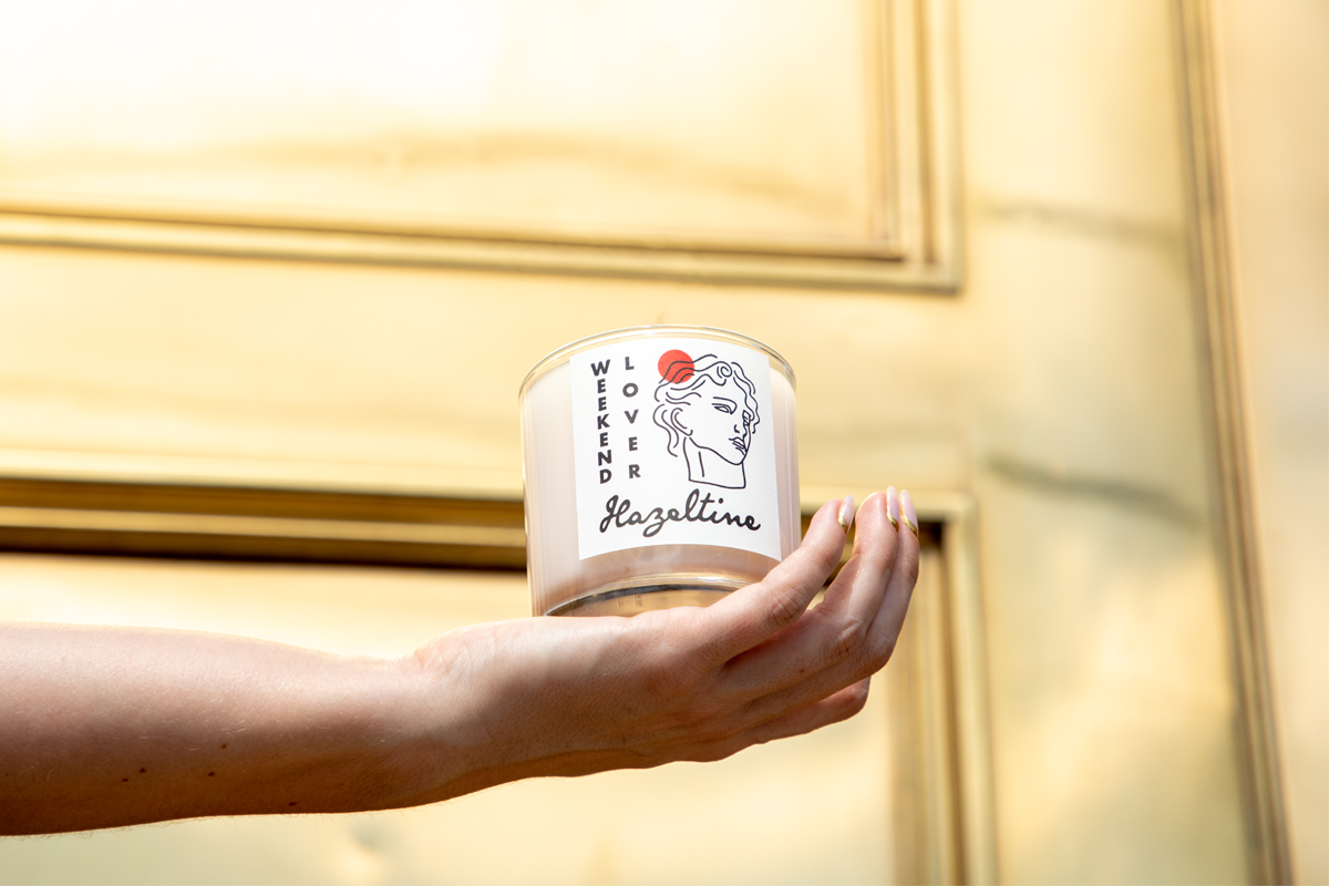 Hand holding a Hazeltine Weekend Lover candle in front of a gold architectural door.
